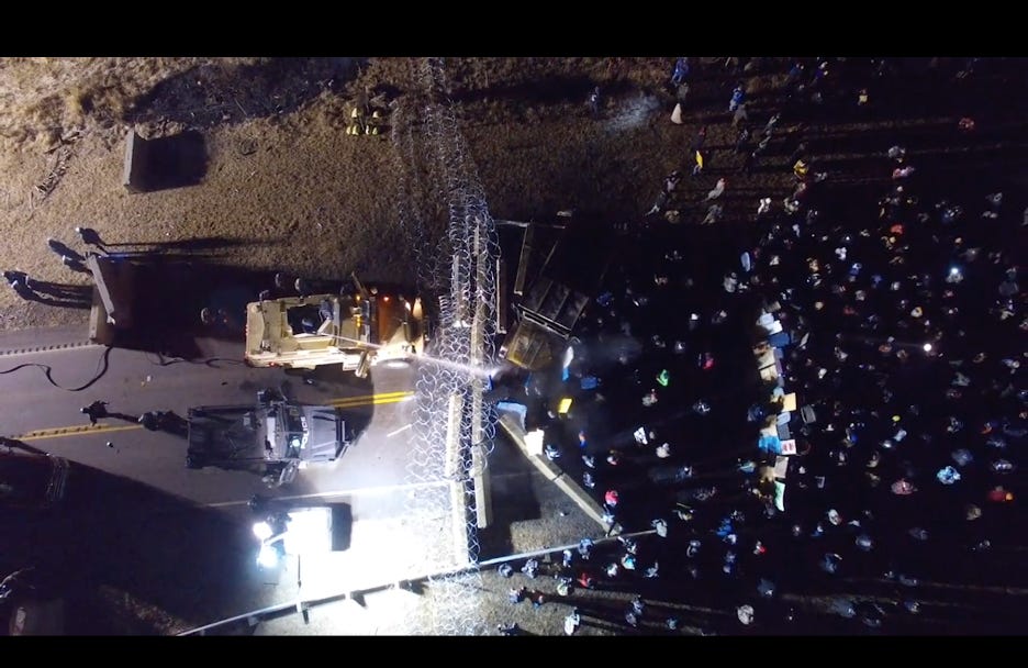 An aerial view of militarized police blocking a road with a barbed wire fence and protesters on the other side.