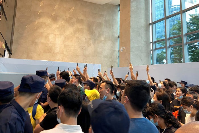A crowd of people gather in the lobby of a building holding up their mobile phones while security guards look on 