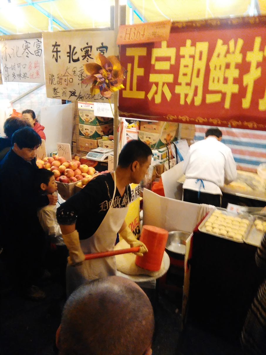 A man in front of a rice cake stall pounding rice cakes by hand with a large red mallet.