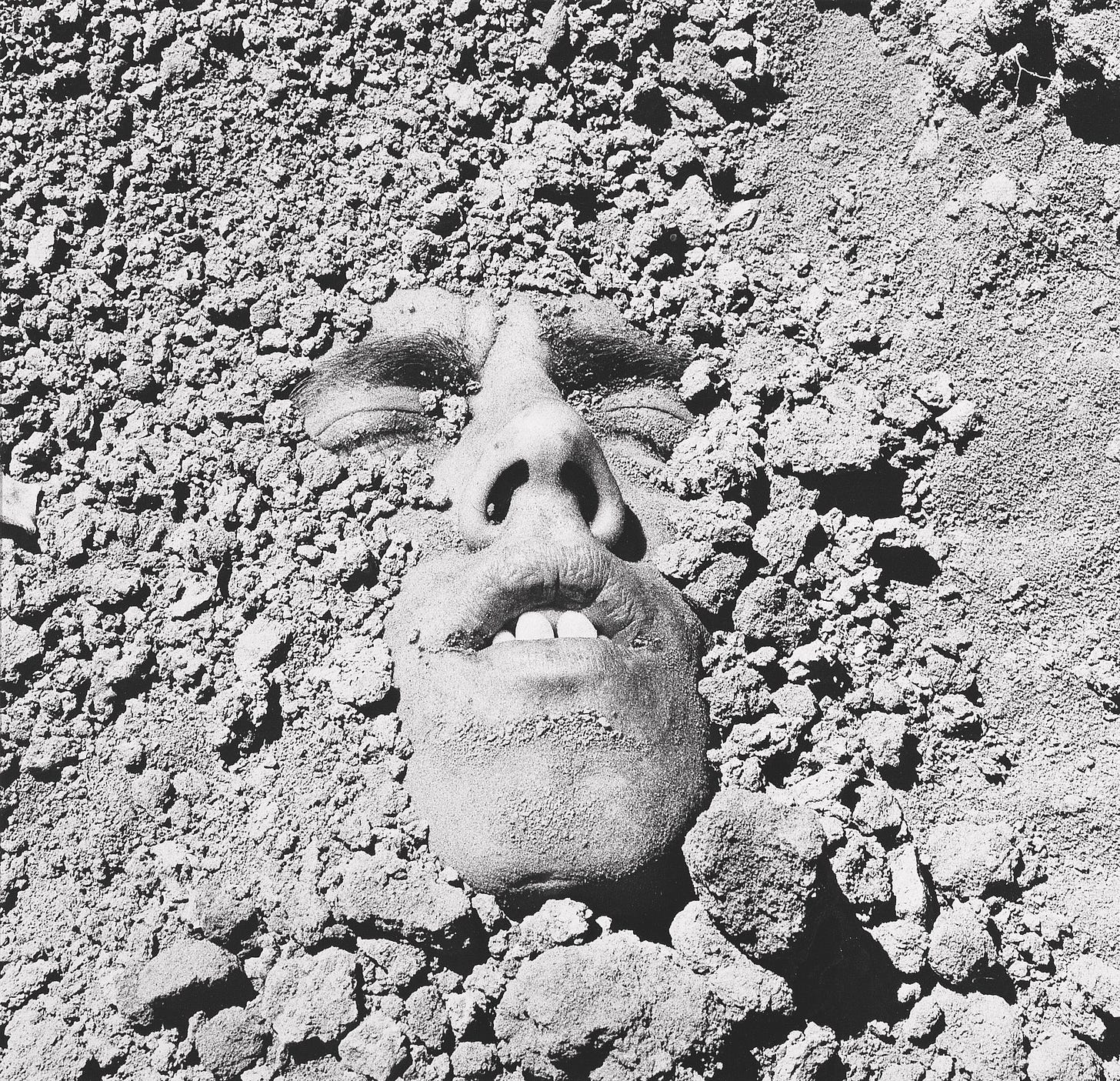 The artist's face is submerged in dirt in a grave.