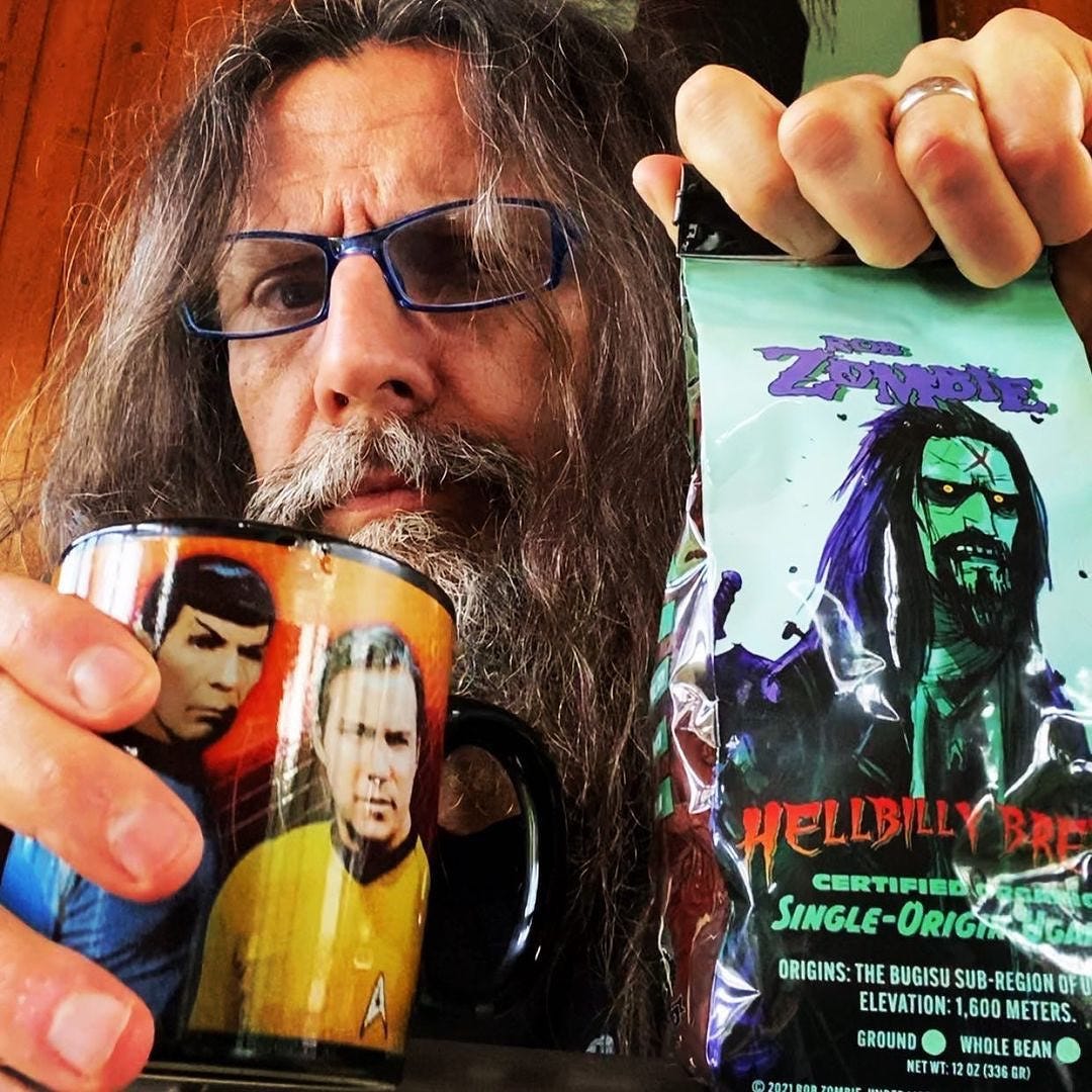 Close up of Rob Zombie holding a cup of coffee and a bag of coffee branded with his name 