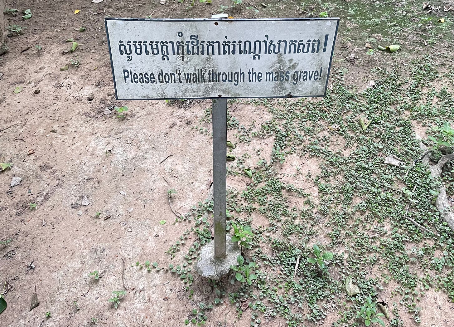 A sign at the Killing Fields that reads "Please don't walk through the mass grave!" in Khmer on top and English on bottom.