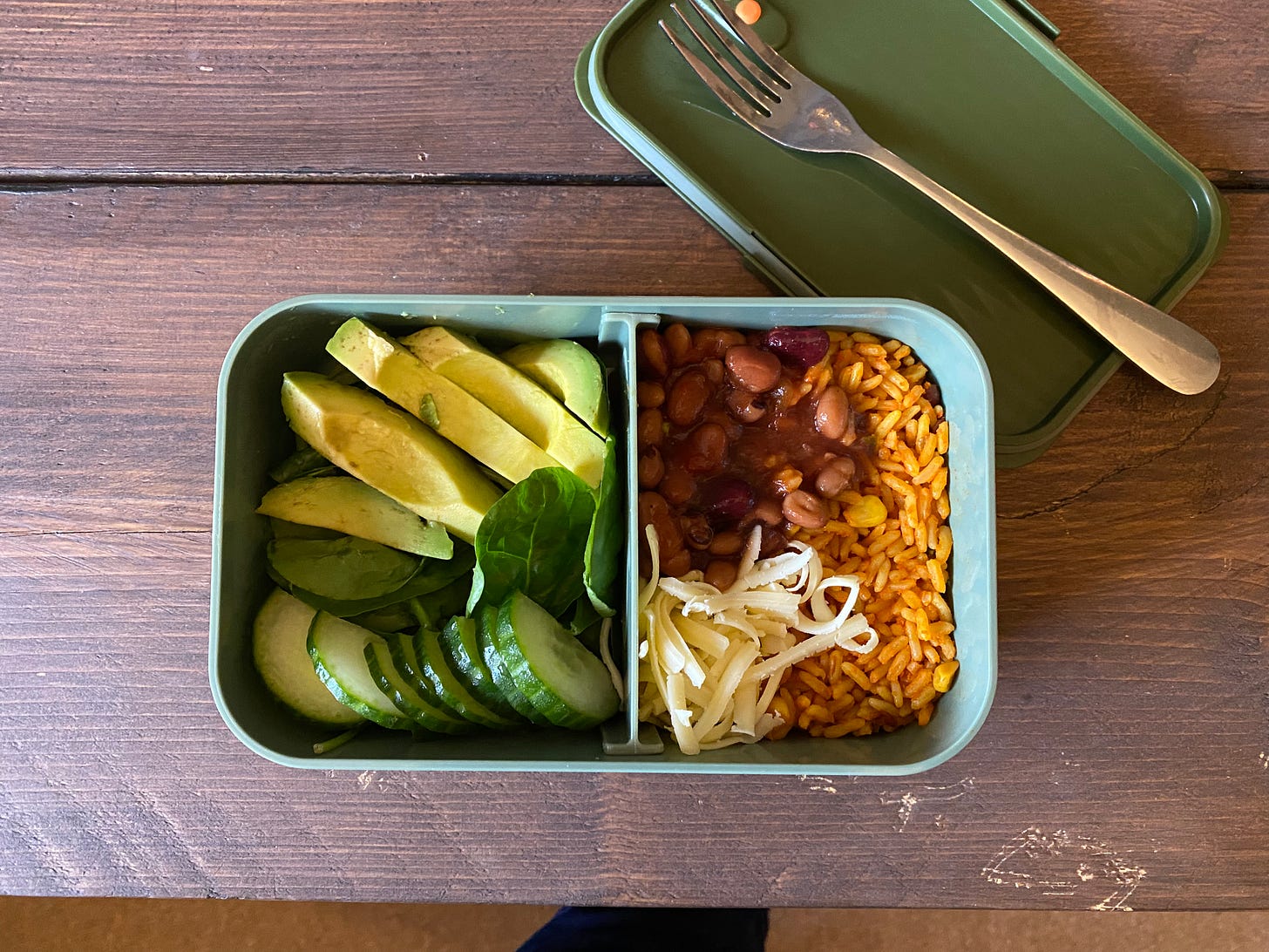 Bento box with lid off, filled with avocado, cucumber, rice, beans and cheese