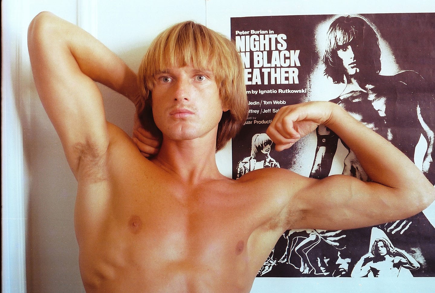 Peter Berlin poses topless next to a poster for his film "Night In Black Leather"
