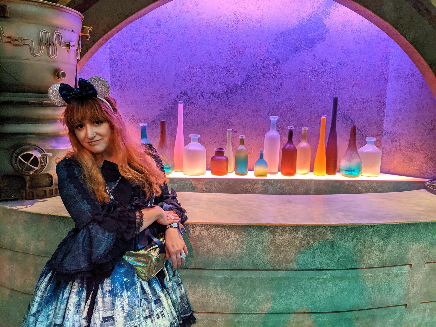 Photo taken in 2019 at Disneyland. Nif (a white femme with red/orange hair) is wearing glittery silver mouse ears with a navy sequined bow. She is wearing Angelic Pretty’s Castle Mirage princess sleeve OP in navy, with a metallic gold fanny pack, and an assortment of AP acrylic jewelry. She is standing in front of a Star Wars bar display, with brightly-colored bottles behind the bar and industrial-styled tank on the bar top.