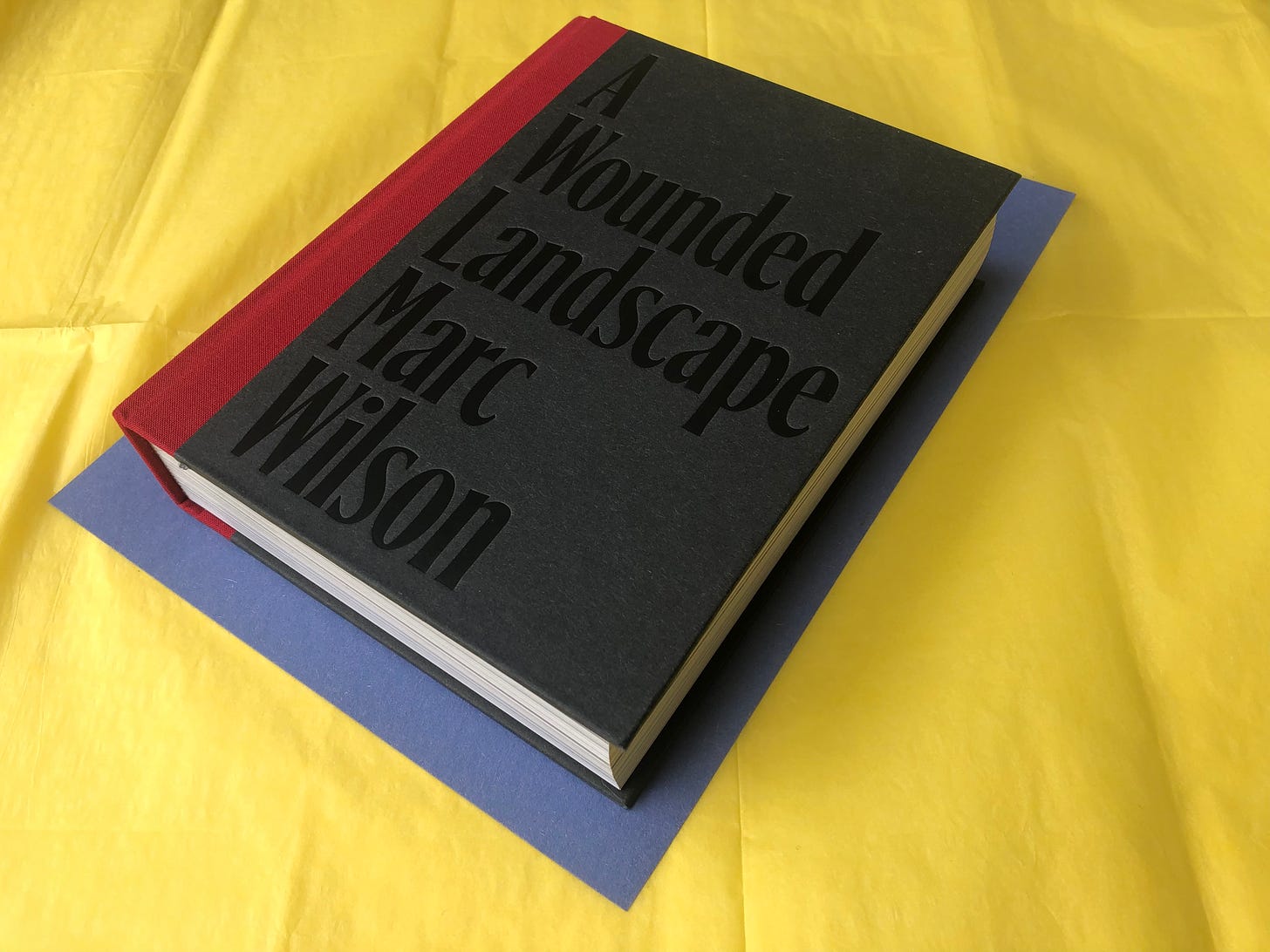 Thick book, red spine, grey cover with A Wounded Landscape Marc Wilson in large letters, each word on a line. Book is on blue paper with yellow tissue underneath