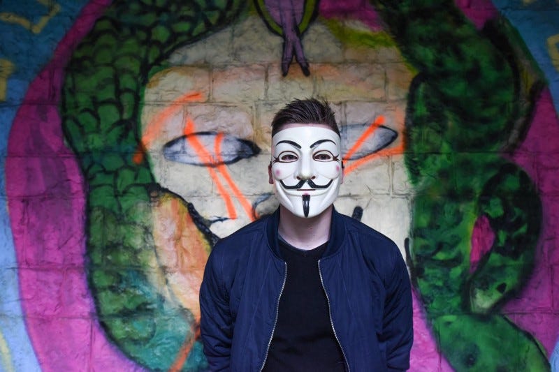 A guy in a Guy Fawkes mask in front of a wall of graffiti