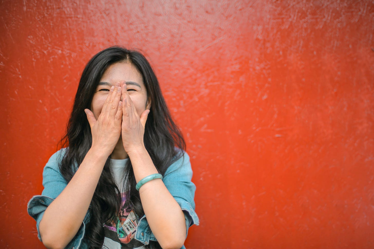 A woman in front of a red wall covering her face with her hands while laughing.