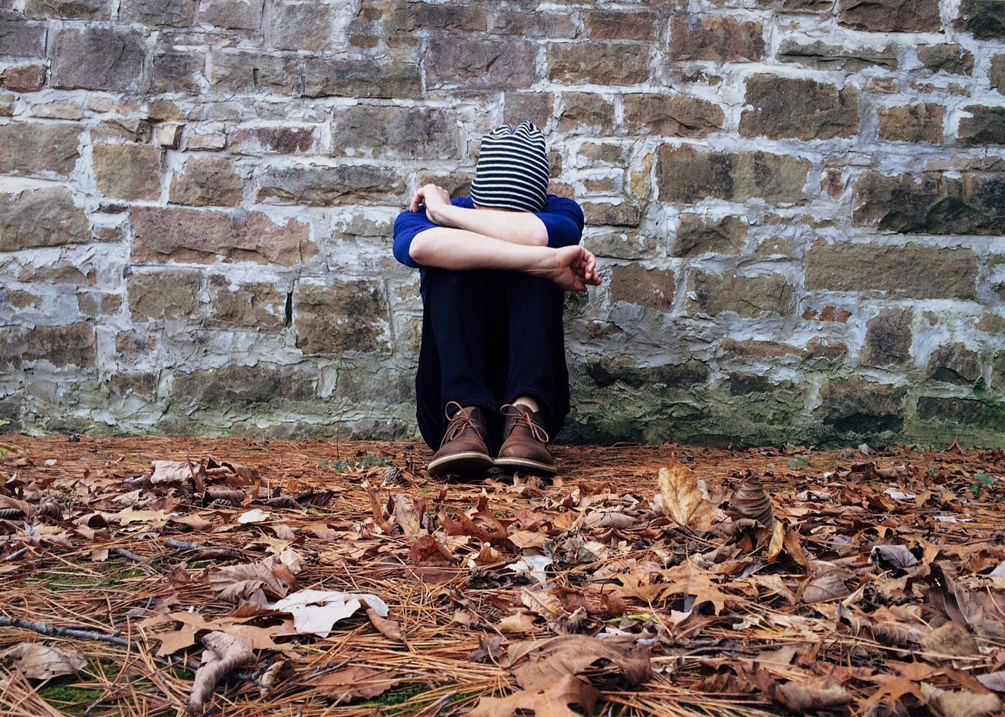 A person sitting against a brick wall while covering their face with their arms.