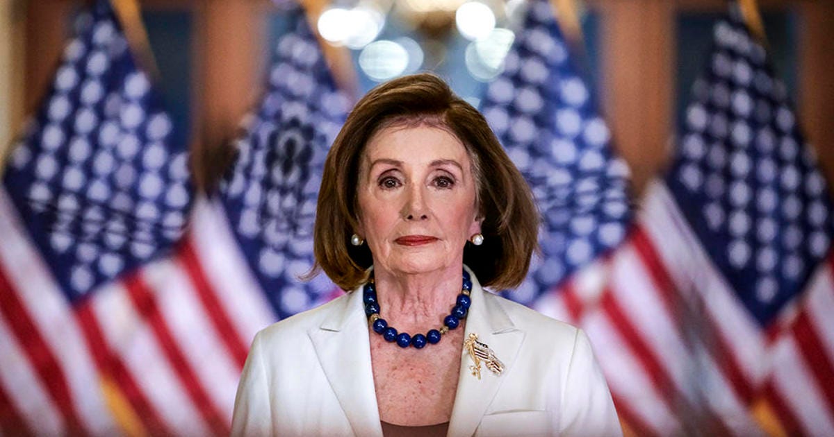 Nancy Pelosi stands in a long hallway, staring forward, beyond the photographer. Behind her, blurry, stand 5 US flags.