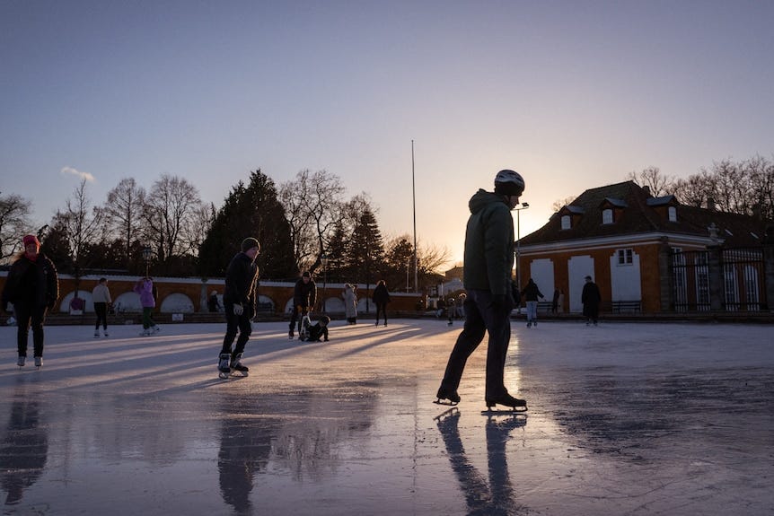 A group of people skate on ice as the sun illuminates their silhouettes behind them.