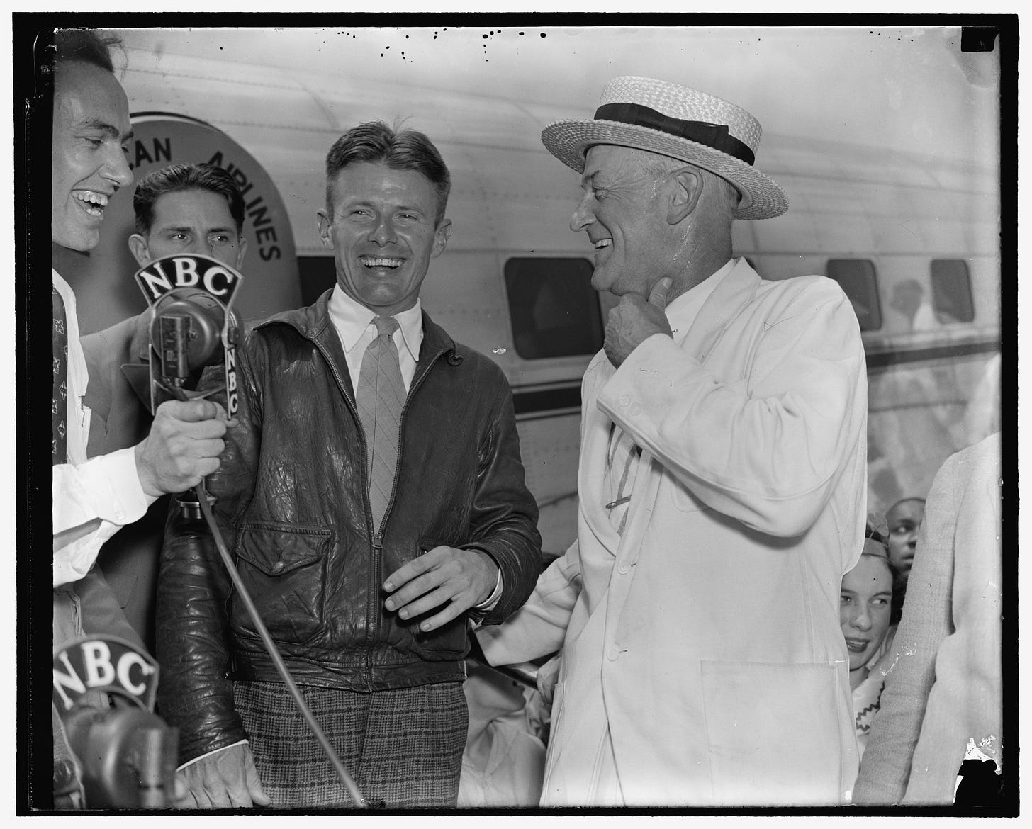 Douglas Corrigan stands before reporters at   an airport. An NBC microphone is visible in the foreground, and an American Airlines Douglas DC-2 is in the background.  Corrigan and the two men standing next to him are laughing.