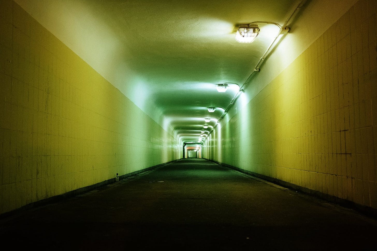 A long hallway or underpass, lit by flourescent lighting. it's the sort of place you wouldn't want to walk down alone. The tiles are white, the grouting is dirty, and there is an eerie glow to the place.