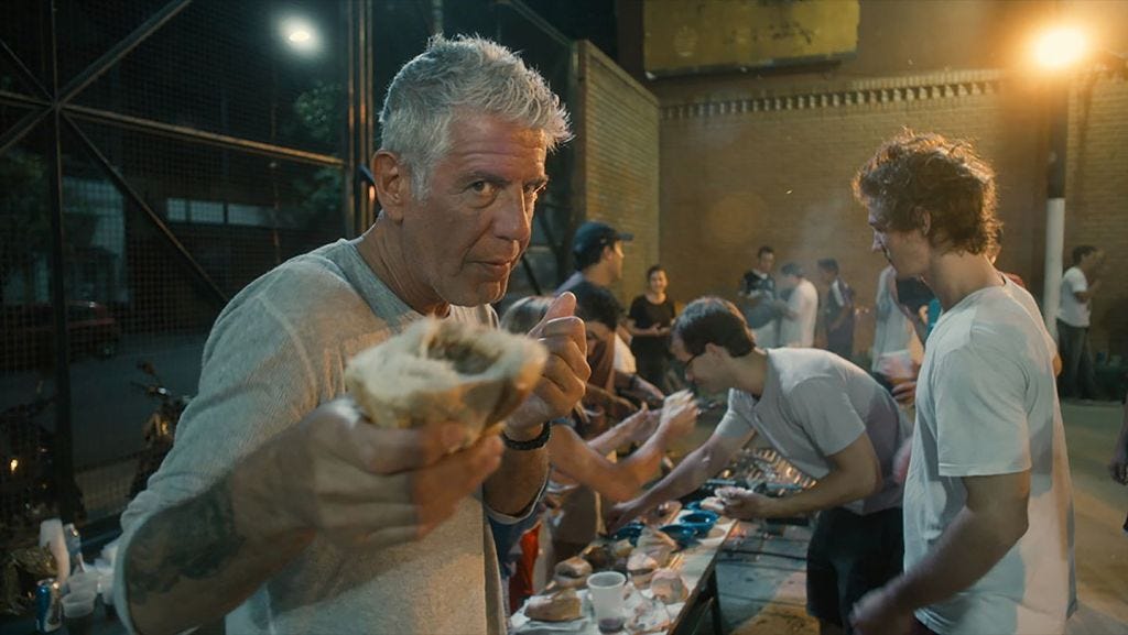 A still from Roadrunner. Anthony Bourdain holds out a sandwich to the camera.