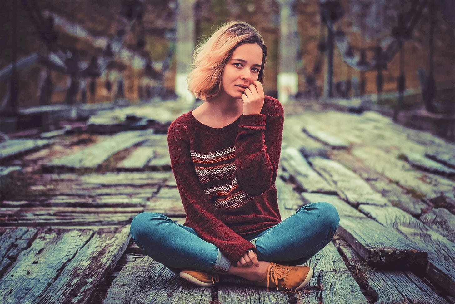 girl wearing long sleeve sweater holding hand to cheek sitting on wooden bridge