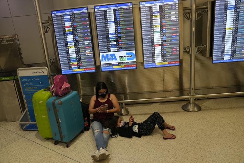 A woman and child from Brazil wait for their flight underneath a board showing roughly a dozen cancelled flights among the scheduled arrivals, at Miami International Airport, Monday, Dec. 27, 2021, in Miami. Thousands of flights worldwide were canceled or delayed on Monday, as airline staffing shortages due to the rapid spread of the omicron variant of COVID-19 continued to disrupt the busy holiday travel season. (AP Photo/Rebecca Blackwell)