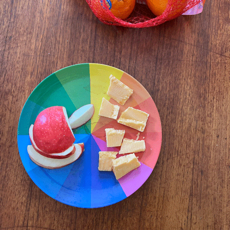 A photo animation of rainbow plate on a table with apple slices and orange chunks of cheddar cheese, with a red plastic mesh bag full of “Cuties” brand oranges in the top right corner and a small hand placing and removing an orange next to the plate.