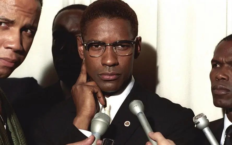 This image shows Denzel Washington as Malcolm X in Spike Lee's 1992 film Malcolm X. Malcolm, with his right hand posed along the right side of his face, stands before three microphones held by offscreen reporters while staring directly into the lens. Malcolm is flanked by three bodyguards.