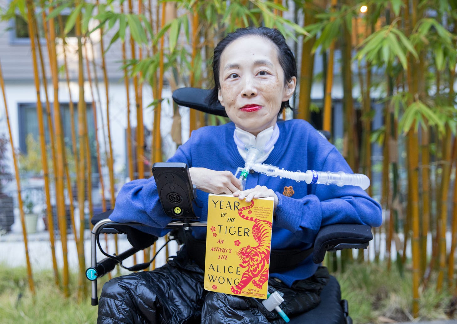 Photo of Alice Wong, an Asian American disabled woman in a power chair. Behind her are bamboo trees. She is wearing a bold red lip color, blue cardigan with a tiny tiger patch, and black puffy pants. A trach and ventilator tube is at her neck and shoulder. She is holding a copy of her memoir, YEAR OF THE TIGER, a yellow book cover with a fierce tiger and red flowers with the text after the title, An Activist’s Life, Alice Wong, Editor of Disability Visibility. Photo credit: Eddie Hernandez Photography.