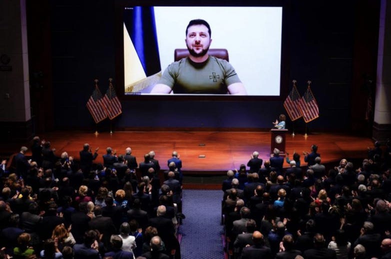 Image description: Ukraine President Zelensky is on a huge screen addressing a huge crowd. On stage on either side of the screen are four US flags, two on either side of the screen. Zelensky himself is wearing an olive green tshirt, sitting in a red leather chair, with a white background behind him and a Ukrainian flag next to him. The audience seated on chairs are all facing forward watching him and cheering.