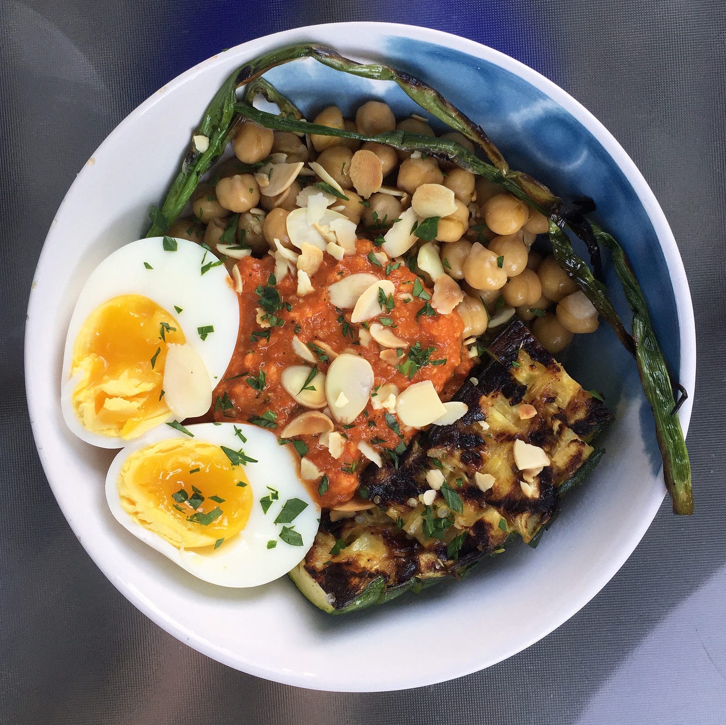 a white bowl with sections of chickpeas, grilled zucchini, and romesco sauce. a soft-boiled egg sliced in half sits to the left, and a charred green onion encircles the top of the bowl. toasted almonds and chopped parsley are scattered on top.