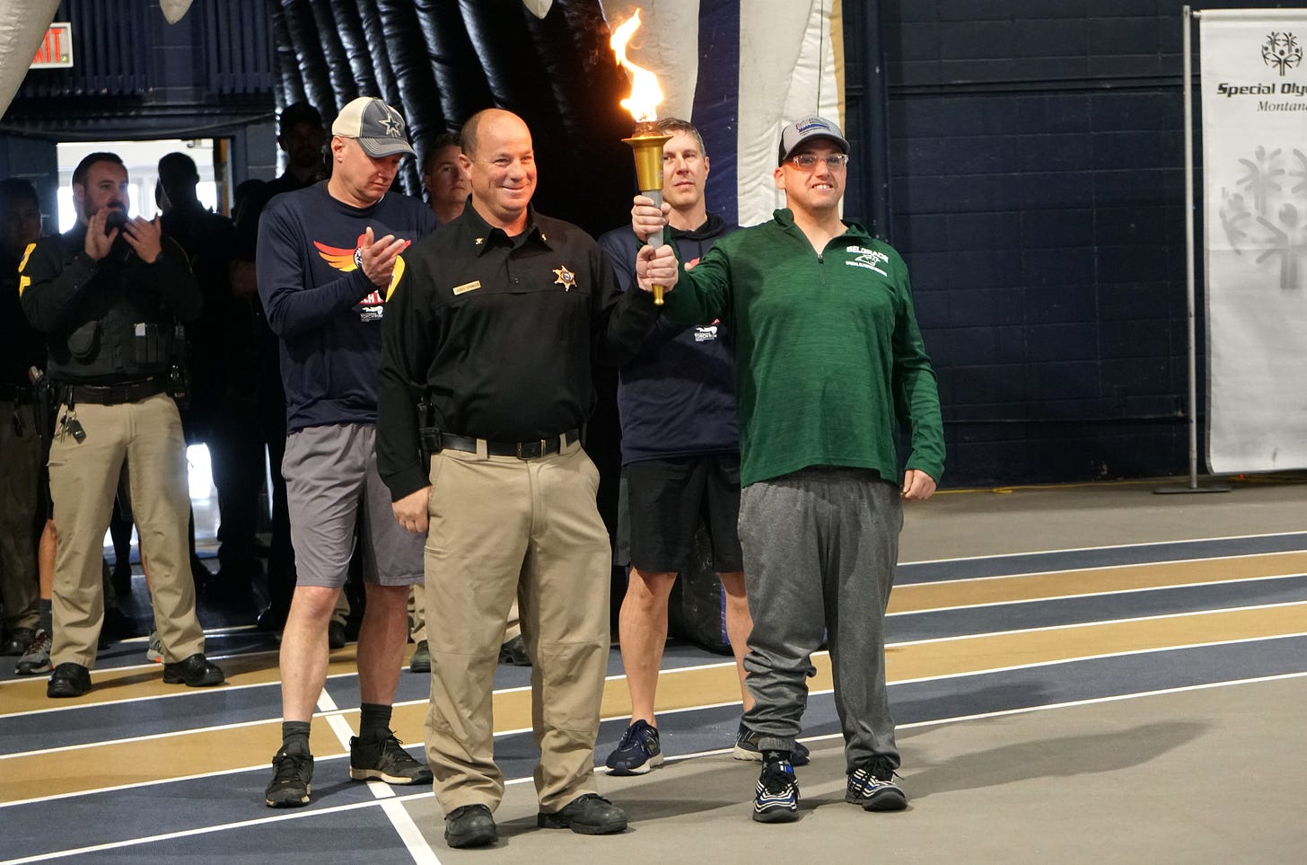 A Special Olympics athlete in a green shirt and gray pants holds the games torch with a law enforcement officer in a black shirt and beige pants and his sheriff’s badge on his chest. Both are smiling. There are several other law enforcement officers behind them smiling and clapping.