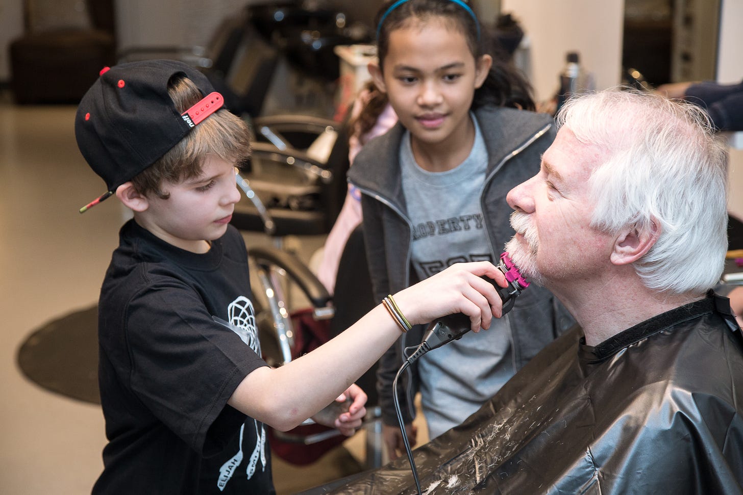 A middle-aged man sits in profile in a salon chair, and two children stand to his front and side, one a boy with electric clippers, gently trimming his silver beard.