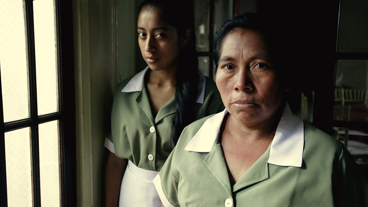 Two indigenous women, one younger and one older, look at the camera, wearing maid uniforms