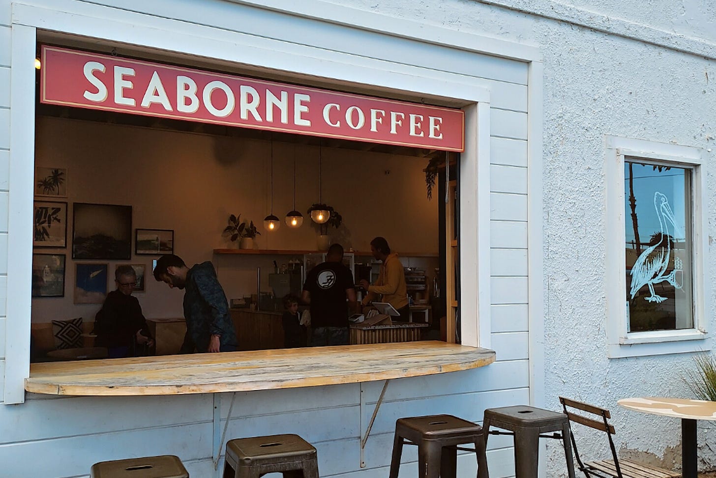 A walk up counter at a coffee shop. Stools outside the white wood and stucco building are set up for customers and an orange sign with white lettering says, "Seaborne Coffee."