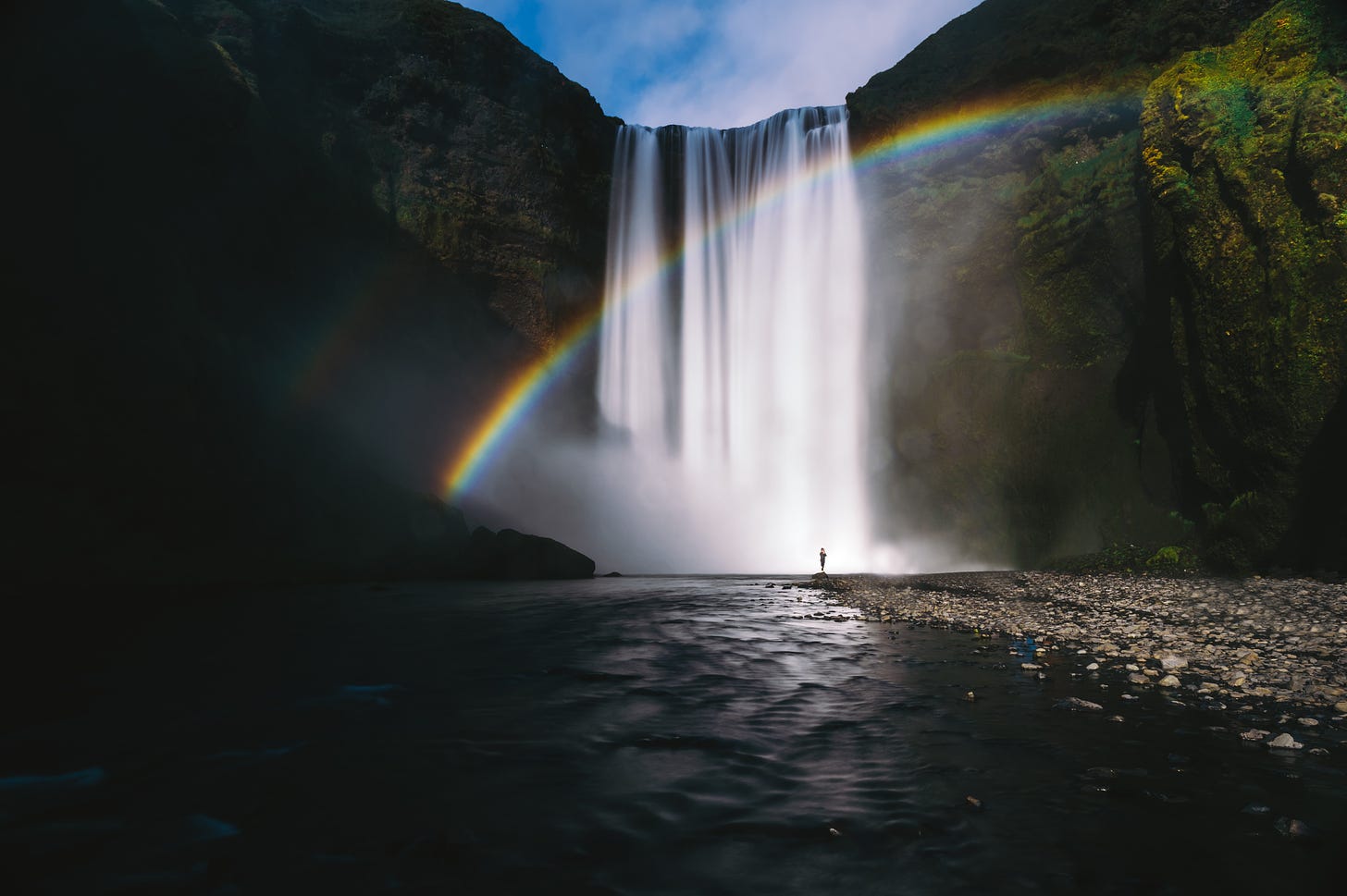 A scenic photo of a waterfall, with a rainbow behind it. A lone figure stands at the bottom of the waterfall.