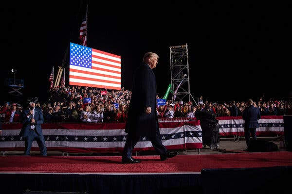 Former President Donald J. Trump walked across a stage surrounded by crowds and a large screen showing an American flag.
