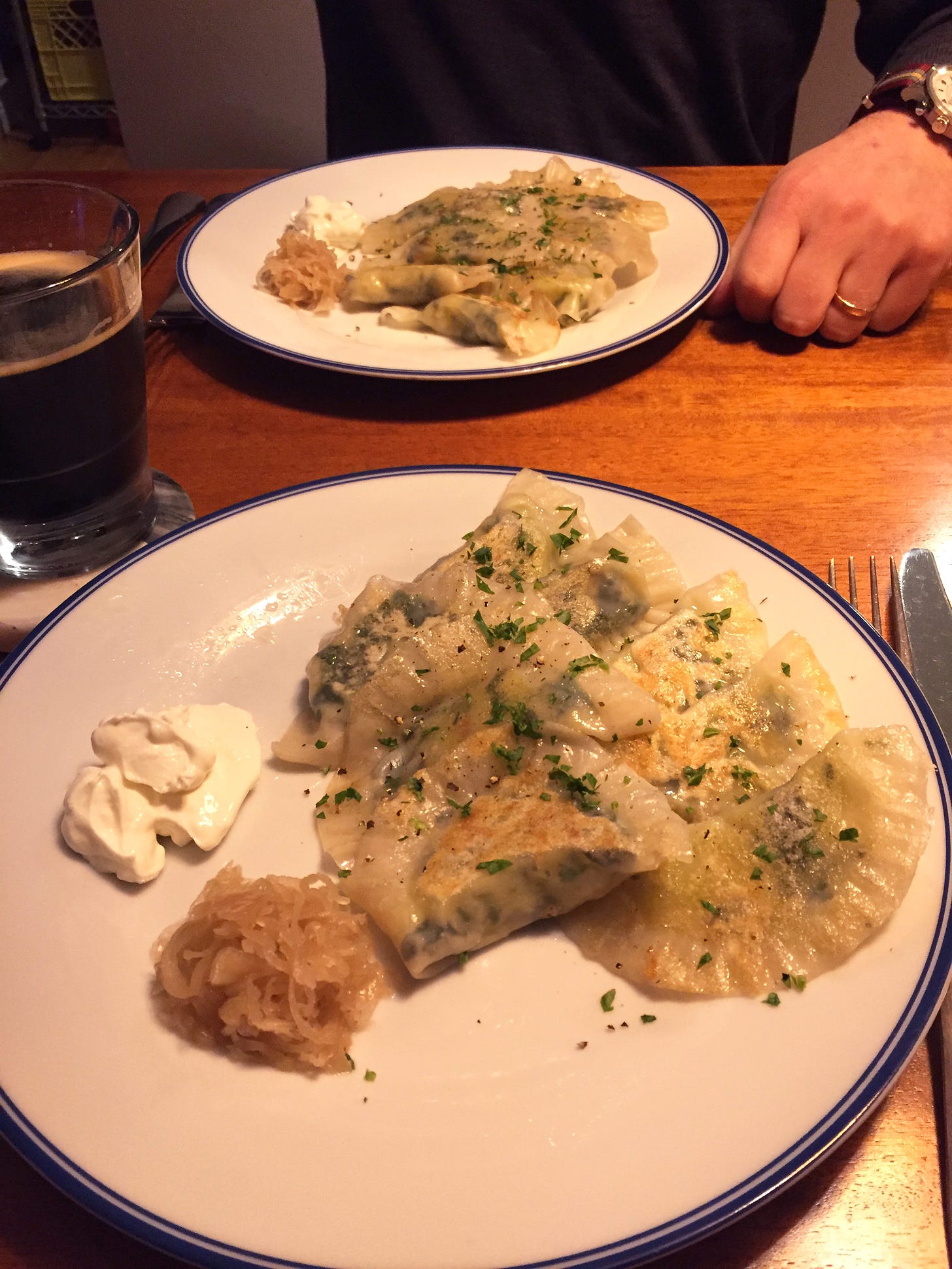 Two plates with eight pierogis arranged in rows of five and three, next to small mounds of sauerkraut and sour cream. A glass of stout sits at the upper left of the plate in the foreground.