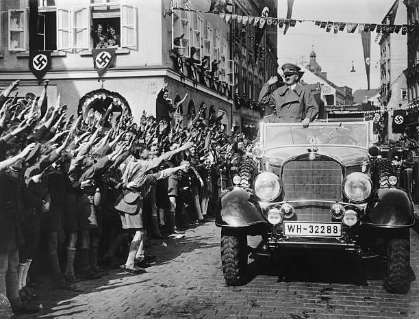 German-speaking schoolchildren welcome German dictator Adolf Hitler in a street decorated with swastikas in the Sudetenland, Czechoslovakia. The...