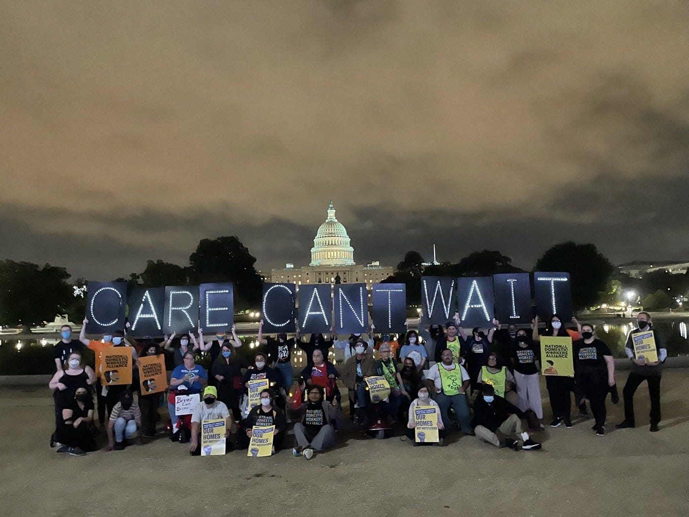About 30 activists gather under a cloudy night sky with the U.S. Capitol building behind them. They carry many signs too small to read, but several signs in large illuminated letters read CARE CANT WAIT.