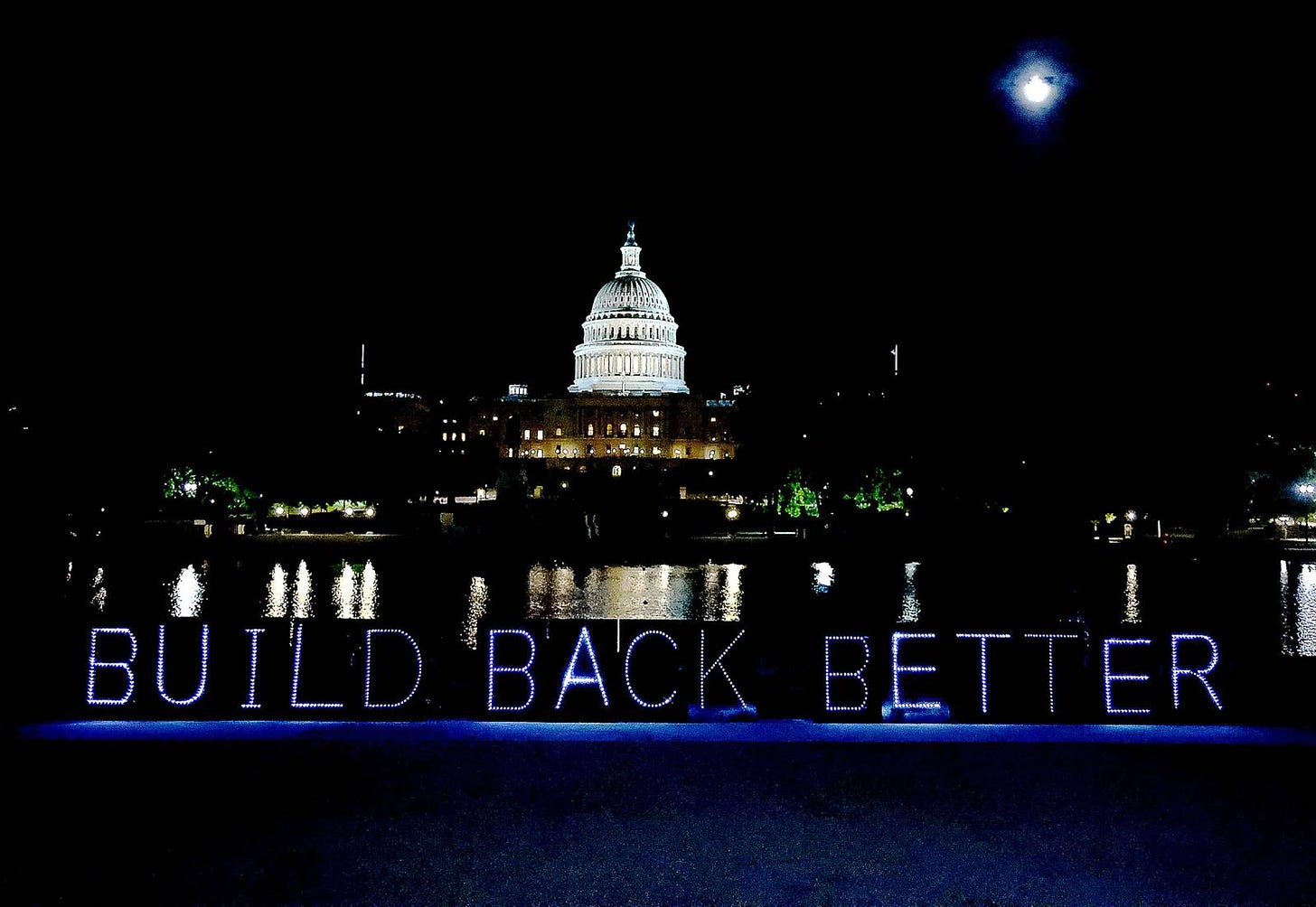At night in front of an illuminated Capitol Building in D.C., light-signs spell out BUILD BACK BETTER.