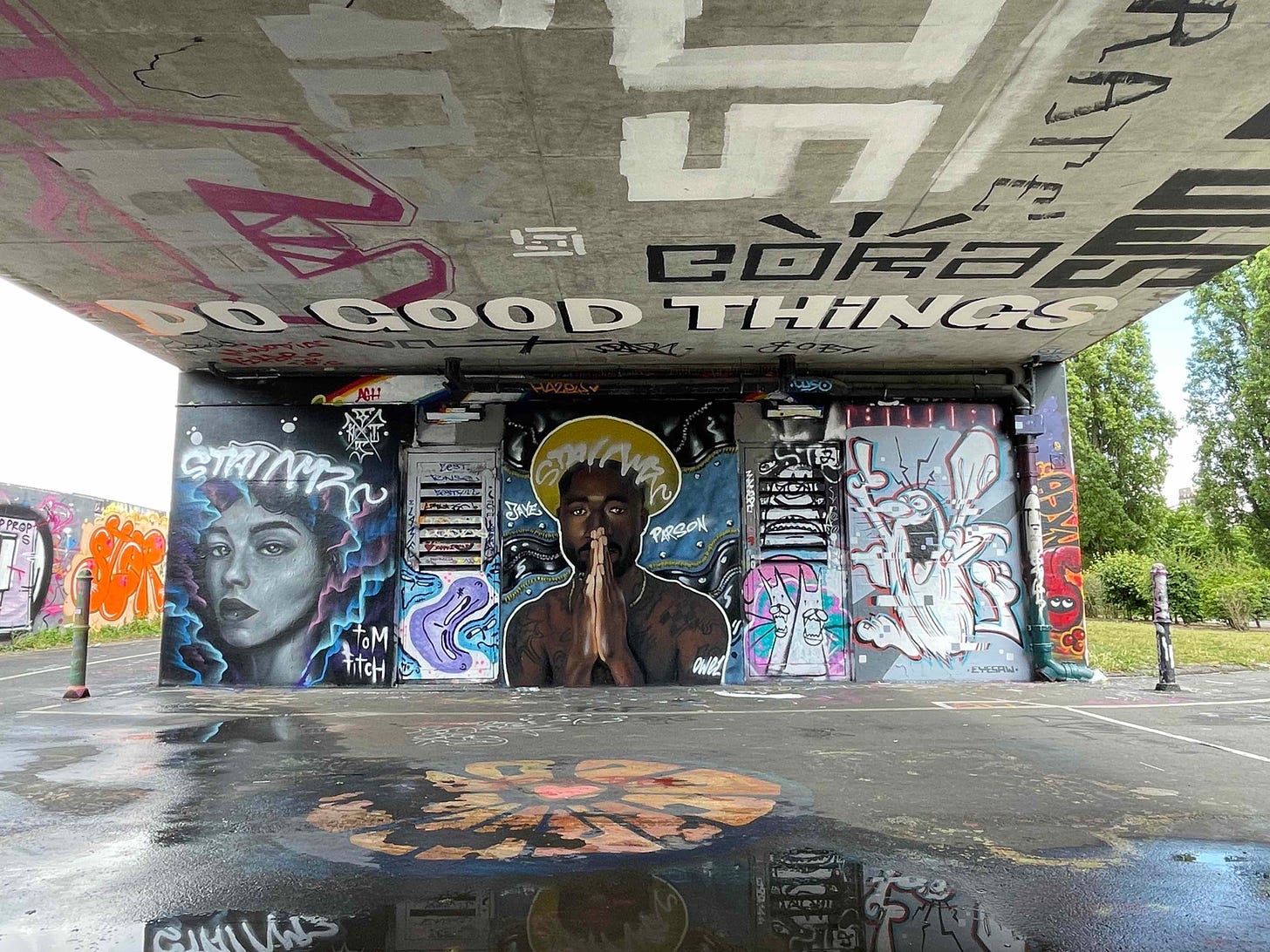 Colour photo showing the wall beneath a railway bridge that is covered in graffiti art depicting a spray painted woman, man and cartoon character, various graffiti tags and the words 'do good things'