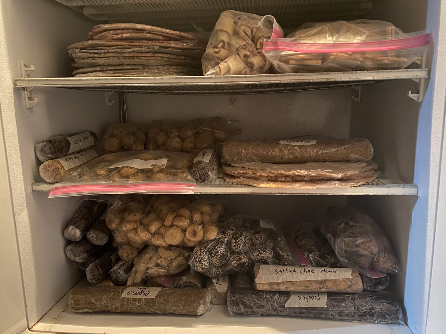 View into a full freezer. Three shelves hold many doughs in the above forms: a stack of rolled out sheets of dough, many dough logs, ziplock of cut out cookies, dough balls, rugelach, and thumbprint cookies.