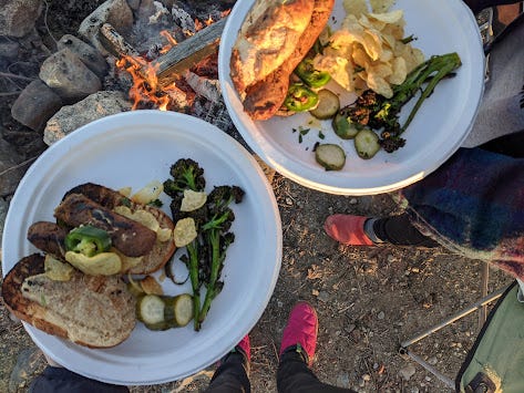 two plates of bratwurst, broccolini and potato chips over a campfire