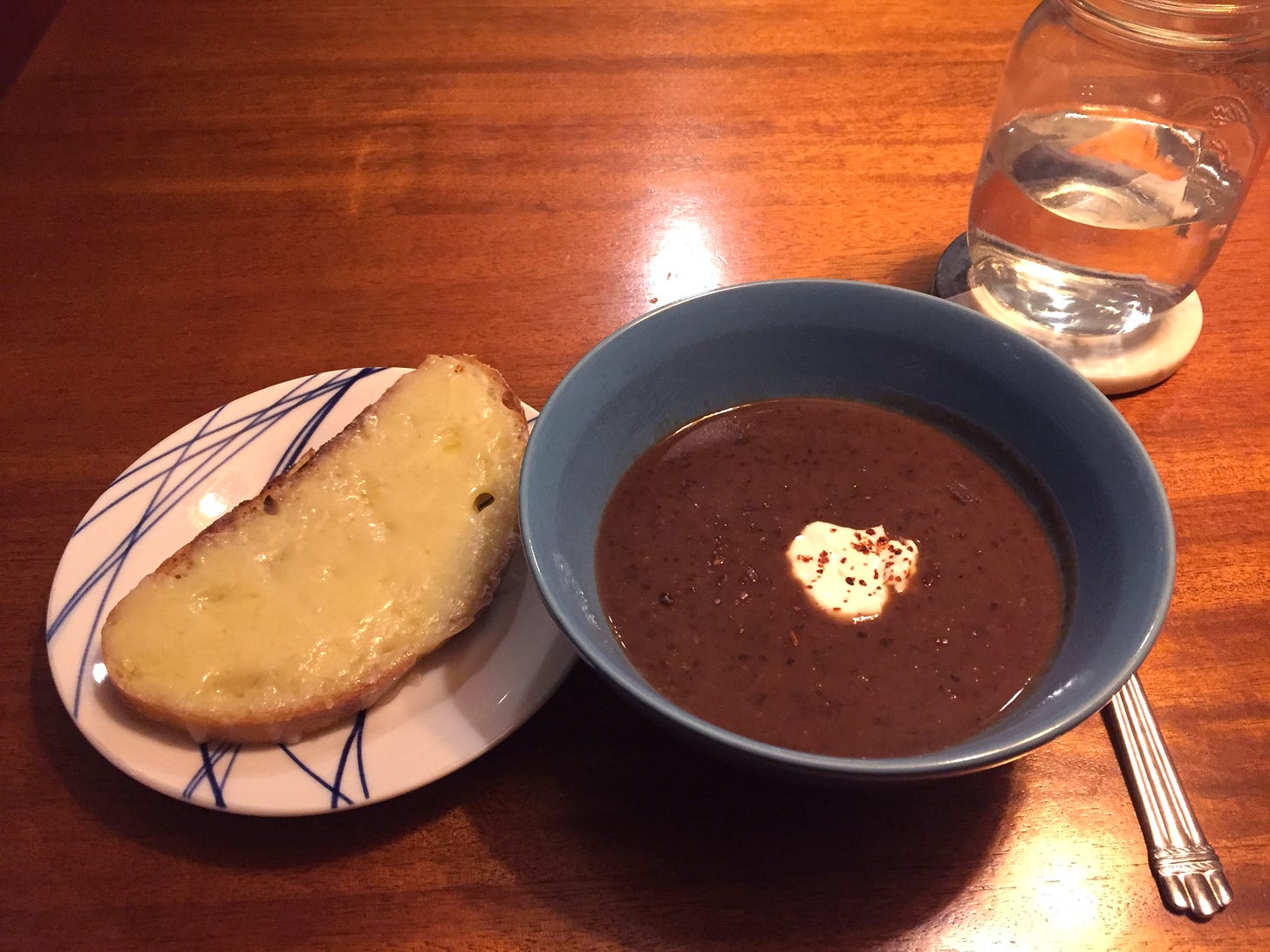 A blue bowl of black bean soup, with chili flakes and a dollop of crema. On a small plate next to it is a slice of sourdough covered in melted white cheddar.
