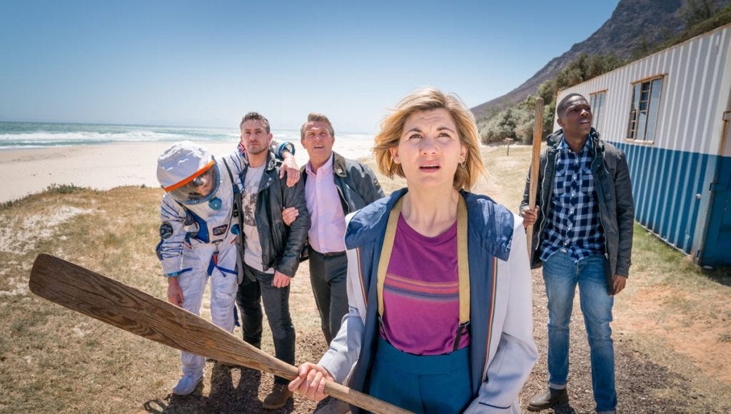 This image shows, from left to right, Adam Lang (played by Matthew McNulty, wearing a European Space Agency astronaut uniform), Jake Willis (played by Warren Brown), Graham O'Brien (played by Bradley Walsh), the Thirteenth Doctor (played by Jodie Whittaker, holding a wooden oar), and Ryan Sinclair (Tosin Cole, also holding an oar) as they look off camera at birds massing in the sky. They stand on a Malagasy beach near a metal laboratory, with the ocean and clear blue sky visible in the background. This image shows, from left to right, Adam Lang (played by Matthew McNulty, wearing a European Space Agency astronaut uniform), Jake Willis (played by Warren Brown), Graham O'Brien (played by Bradley Walsh), the Thirteenth Doctor (played by Jodie Whittaker, holding a wooden oar), and Ryan Sinclair (Tosin Cole, also holding an oar) as they look off camera at birds massing in the sky. They stand on a Malagasy beach near a metal laboratory, with the ocean and clear blue sky visible in the background.
