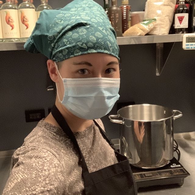 A woman wearing a surgical mask, apron, and bandana in front of a large pot