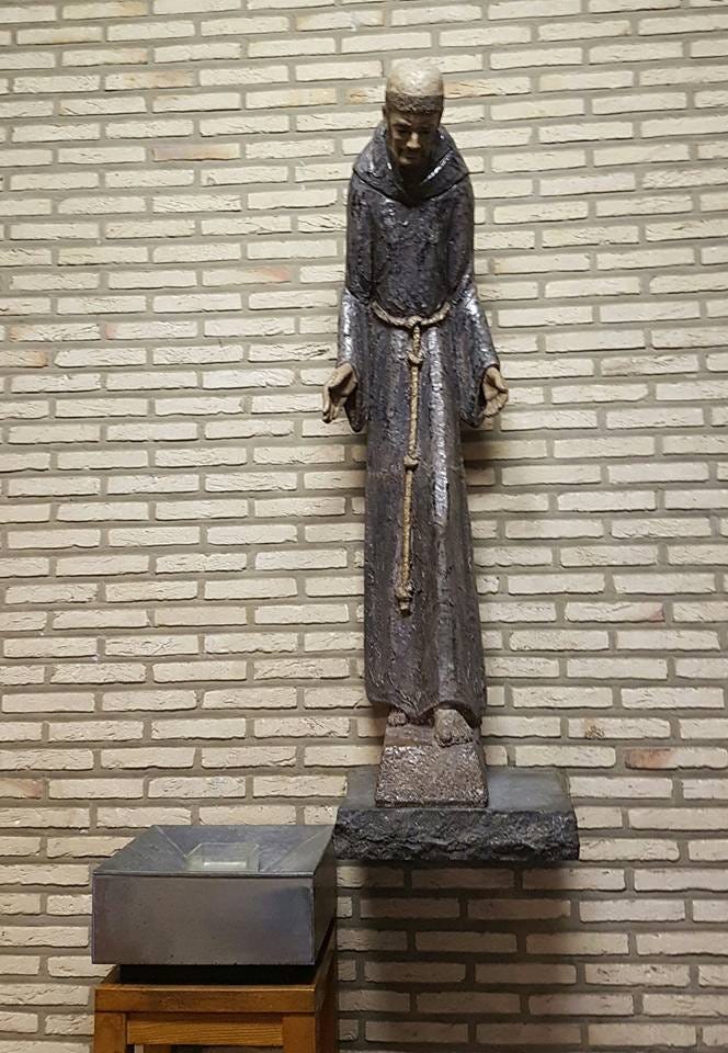 En keramikstaty av en väldigt mager man med tonsur i brun ordensdräkt. Bakom statyn syns en ljus tegelvägg.