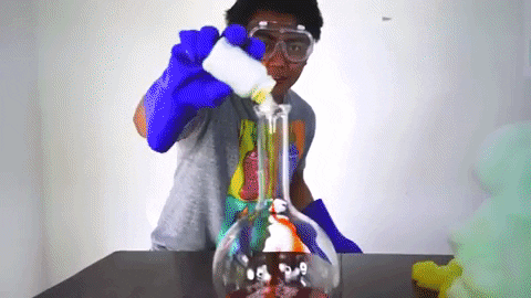 A young man wearing blue rubber gloves dumps a substance into a flask and orange foam shoots up to the ceiling