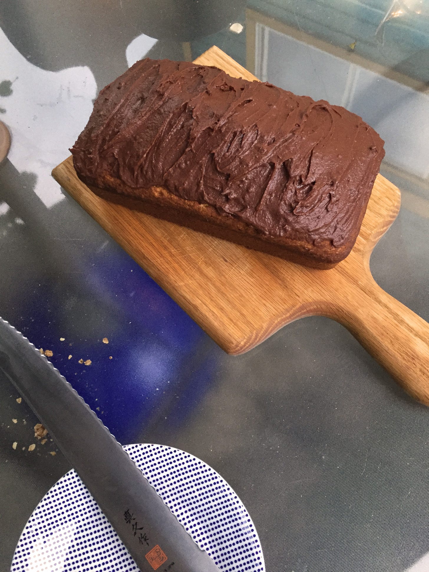 On a small wooden bread board, a loaf of banana bread with swirls of chocolate ganache spread on top. A bread knife sits on the table to its lower left.
