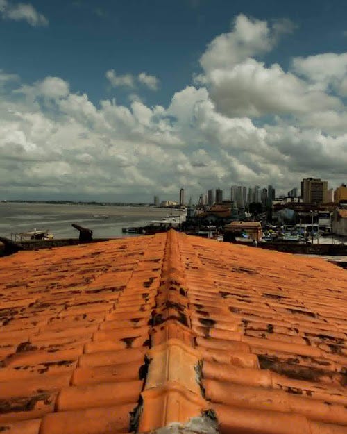 Fotografia do horizonte de Belém, Pará. Na parte superior, o céu azul com algumas nuvens. No centro, a esquerda, o rio, e a direita, os prédios da cidade. Na parte inferior, as telhas do telhado, de onde a foto está sendo tirada. A imagem é vertical.