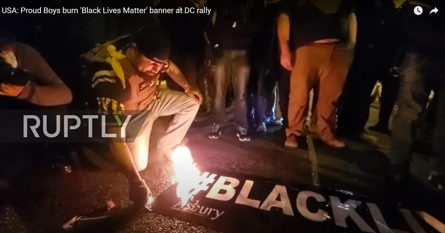A screenshot of a YouTube video, title in corner: USA: Proud Boys burn 'Black Lives Matter' banner at DC rally. On camera, a heavyset man with a brown beard, a backwards baseball cap, a plate carrier, and Proud Boys colors on his shirt kneels to light a Black Lives Matter banner on fire. He is very clearly not Enrique Tarrio. Many people surround the banner