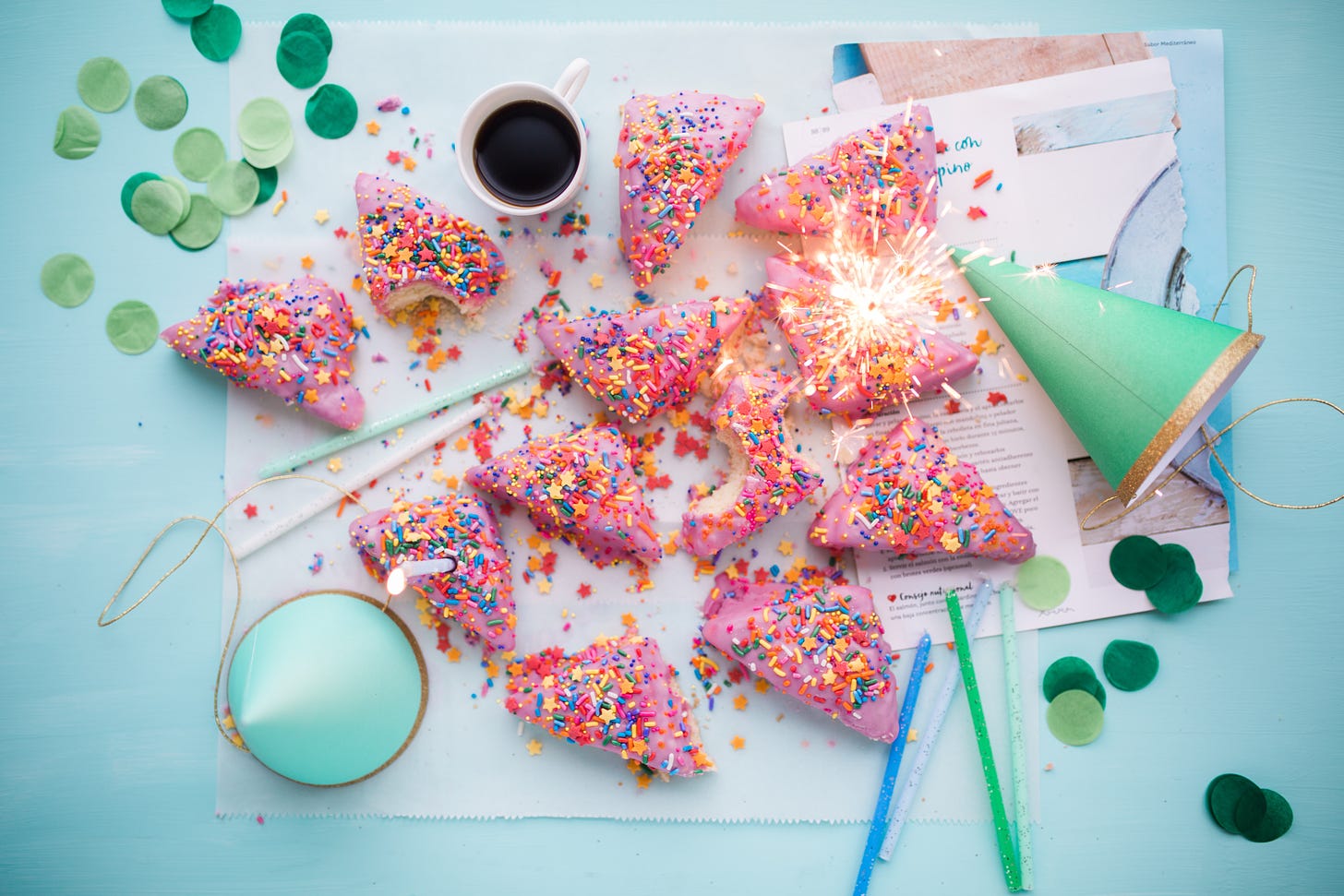 A party mess with triangular pink cupcakes with sprinkles on a table, a couple of party hats, a cup of coffee, some candles, and some paper circle decorations.