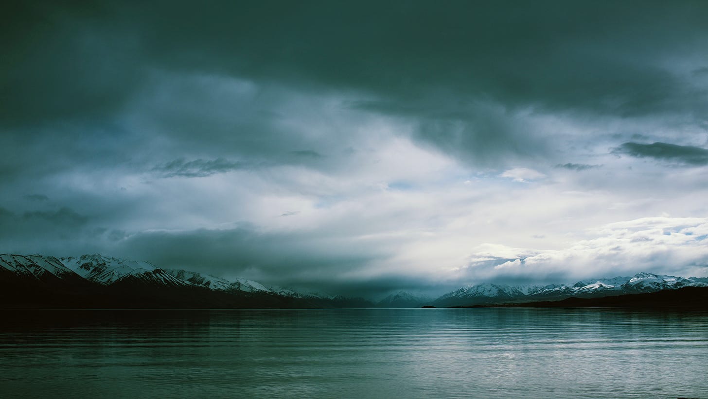 Photograph of Lake Pukaki under a cloudy sky looking towards snowcapped mountains. The lake is still with a few ripples. 