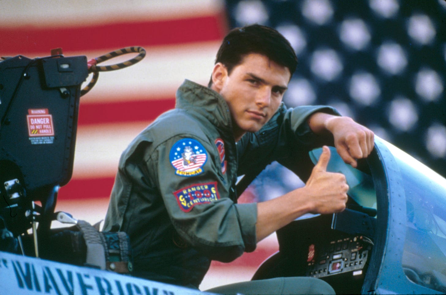 An image of actor Tom Cruise in the cockpit of an F-16, making a thumbs up gesture.