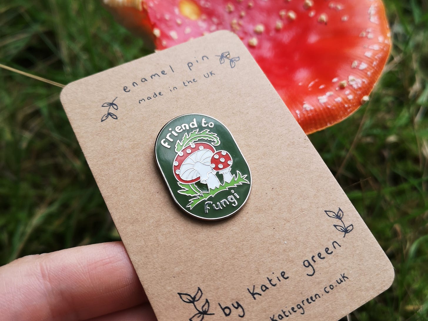 Image description: a white person's hand holding a brown card bearing a cute shiny enamel pin. It's oval shaped with a dark green background. The design shows two cute fly agaric mushrooms snuggling up together with a frond of bracken arching over them. The text in silver says Friend to Fungi. In the background is a real fly agaric mushroom.